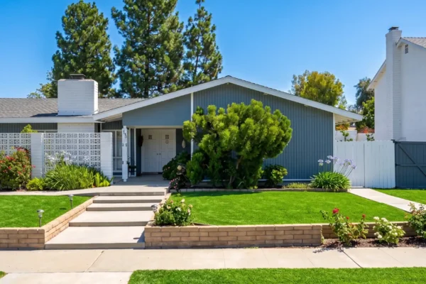 "A perfectly centered, straight-on architectural photograph of a single-story home with light blue siding and white trim. The composition is flat and highly symmetrical, dividing the frame into a clear, vibrant blue sky at the top, the strong horizontal lines of the house in the midground, and a bright, manicured green lawn in the foreground. Bright, natural daylight illuminates the scene, casting soft, warm shadows from the bushes and roof eaves that add depth to the facade. The vertical house number '1482' is visible on the white pillar next to the recessed front entrance."