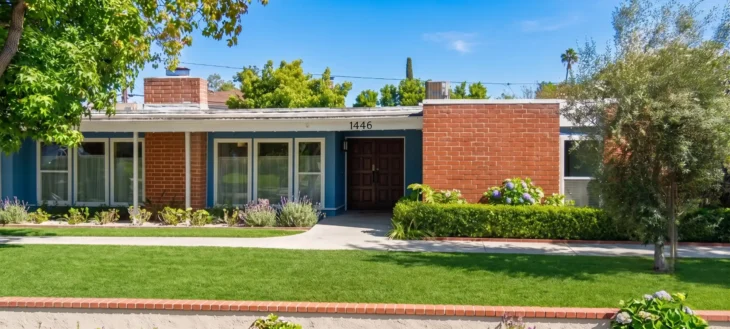 A bright, wide-angle photograph captures a mid-century modern house facade under a clear blue sky. The scene is illuminated by strong, warm sunlight, casting defined, crisp shadows. The house is a long, low, single-story structure, asymmetrical in design. Its walls are composed of horizontal siding in a bright blue-gray color and large sections of textured reddish-brown brickwork. The central focus is dark wooden double doors with recessed panels, above which the numbers '1446' are clearly visible in white. Large, white-framed windows with vertical blinds line the left and right sections. Llandscaping includes a meticulous foreground lawn, a clean concrete walkway, a low brick border, and layered plantings of flowering lavender and blue hydrangeas. A large mature tree with dense green leaves flanks the left side, and a smaller, more open olive tree is to the right. The entire composition has visible Google Maps UI elements, including a small map overlay with the text 'ACACIA Elementary School' in the bottom left, and navigation tools in the bottom right, overlaid on the grass and foliage.