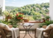Bright editorial photography of a sunlit apartment balcony bathed in soft, natural morning light with a clean, centered composition. Two woven wicker armchairs with cream cushions and light throws flank a small bistro table holding an open crossword book and a coffee cup. Vibrant red geraniums and leafy potted plants line the terracotta railing, framing a crisp view of a lush green hillside in the distance under a clear blue sky, evoking a warm, inviting, and peaceful atmosphere.