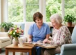 A brightly lit, photorealistic composition bathed in soft, warm natural sunlight streaming through large background windows. The image features a shallow depth of field, gently blurring the green foliage outside and drawing focus to the center. Vibrant, true-to-life colors are highlighted by a vase of fresh spring flowers in the foreground. Two figures, bathed in soft daylight, are seated in plush armchairs, leaning over a wooden table to view an open photo album. The overall tone is airy, clean, and inviting. No text is visible.