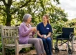 A wide-angle, medium shot bathed in warm, natural sunlight, depicting a senior woman and a younger caregiver seated side-by-side on a weathered wooden bench. The elderly woman in a purple cardigan and the caregiver in blue scrubs hold ceramic mugs, smiling while engaged in conversation. An empty wheelchair is positioned to the right. The background is a lush garden with blooming hydrangeas and large oak trees under a partly cloudy sky, featuring dappled light on the foliage. The caregiver's uniform has a small, illegible white name tag.